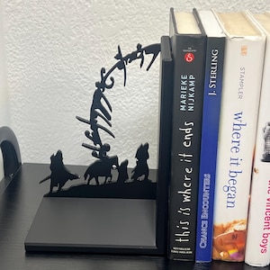 Op de afbeelding: Zwarte metalen boekensteunen met een silhouetontwerp van figuren te paard en lopend. De boekensteunen houden verschillende boeken vast, waaronder "This is Where it Ends" en "Chance Encounters".