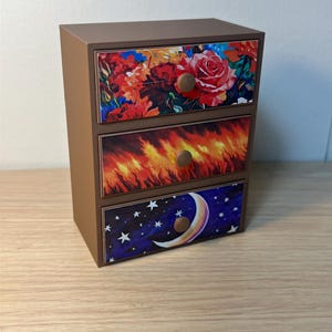 Puede incluir: Un peque&ntilde;o cofre marr&oacute;n de tres cajones con frentes decorativos. El caj&oacute;n superior presenta un dise&ntilde;o floral con rosas rojas. El caj&oacute;n central tiene un dise&ntilde;o de llama ardiente, y el caj&oacute;n inferior muestra un cielo nocturno con una luna creciente y estrellas.