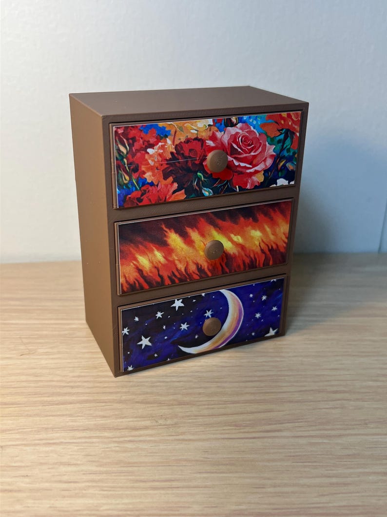 Puede incluir: Un peque&ntilde;o cofre marr&oacute;n de tres cajones con frentes decorativos. El caj&oacute;n superior presenta un dise&ntilde;o floral con rosas rojas. El caj&oacute;n central tiene un dise&ntilde;o de llama ardiente, y el caj&oacute;n inferior muestra un cielo nocturno con una luna creciente y estrellas.