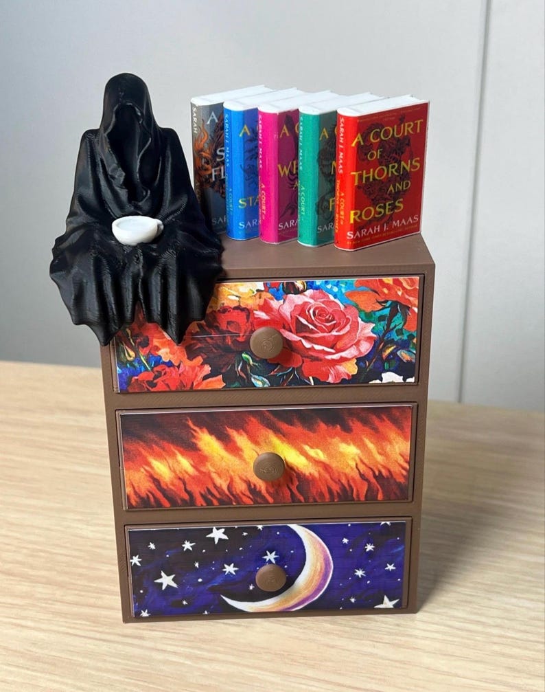 Puede incluir: Un cofre en miniatura de tres cajones con dise&ntilde;os florales, de fuego y celestiales. Una figura negra con capucha est&aacute; sentada en el cofre, sosteniendo una taza blanca. Peque&ntilde;os libros con lomos coloridos est&aacute;n dispuestos encima. Los t&iacute;tulos de los libros incluyen "A Court of Thorns and Roses".