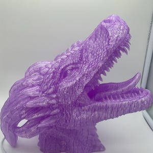 Puede incluir: Una detallada escultura de cabeza de dragón impresa en 3D en un vibrante tono púrpura. La boca del dragón está abierta, revelando afilados dientes y una lengua texturizada. La escultura presenta escamas intrincadas y un ojo detallado.