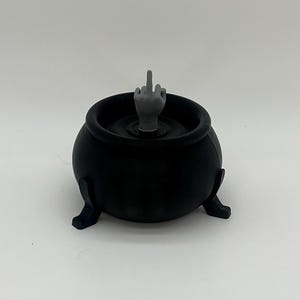 Puede incluir: Un caldero negro con tres patas, con una mano gris que muestra el dedo medio que sale del l&iacute;quido en su interior. El caldero es un art&iacute;culo de novedad, probablemente para Halloween o un tema similar.