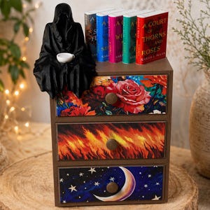 Puede incluir: Un cofre de madera decorativo de tres cajones con dise&ntilde;os florales, de fuego y celestiales. Una figura negra se sienta encima del cofre, y se exhiben varios libros. El cofre est&aacute; sobre una superficie de madera, con un jarr&oacute;n y una vela en el fondo.