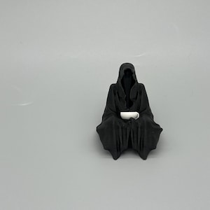 Puede incluir: Una figura negra de una figura con capucha y capa, sentada y sosteniendo una taza blanca. La figura tiene una apariencia oscura y sombreada, con la taza que aparece brillante contra el negro. La figura est&aacute; sobre un fondo gris liso.