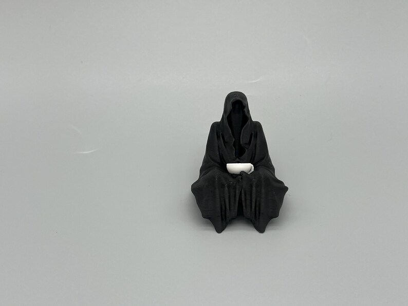 Puede incluir: Una figura negra de una figura con capucha y capa, sentada y sosteniendo una taza blanca. La figura tiene una apariencia oscura y sombreada, con la taza que aparece brillante contra el negro. La figura est&aacute; sobre un fondo gris liso.