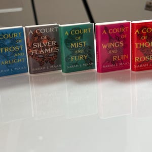 Puede incluir: Una colecci&oacute;n de cinco libros en miniatura de la serie "A Court of Thorns and Roses" de Sarah J. Maas. Los libros tienen cubiertas coloridas en tonos azul, plateado, verde, rosa y rojo, con dise&ntilde;os intrincados y t&iacute;tulos.