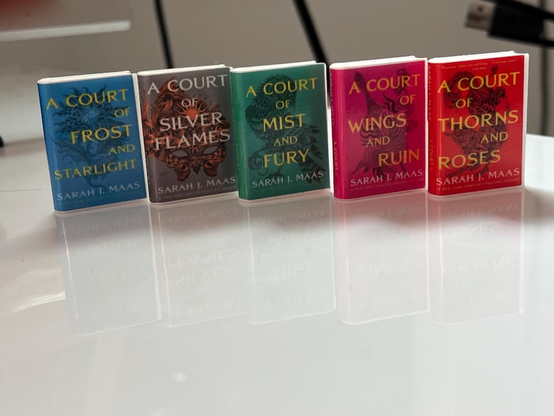 Puede incluir: Una colecci&oacute;n de cinco libros en miniatura de la serie "A Court of Thorns and Roses" de Sarah J. Maas. Los libros tienen cubiertas coloridas en tonos azul, plateado, verde, rosa y rojo, con dise&ntilde;os intrincados y t&iacute;tulos.
