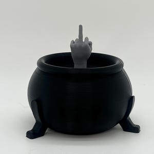 Puede incluir: Un caldero negro con tres patas, con una mano gris haciendo una peineta. El caldero es un objeto redondo tridimensional, probablemente hecho de pl&aacute;stico o resina, con un acabado mate.