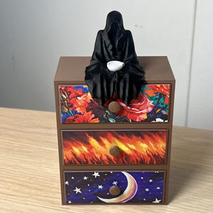 Puede incluir: Un cofre decorativo marr&oacute;n de tres cajones con frentes de cajones florales, de fuego y celestiales. Una figura negra con capucha con un cuenco blanco se sienta encima del cofre. Los cajones tienen pomos marrones.