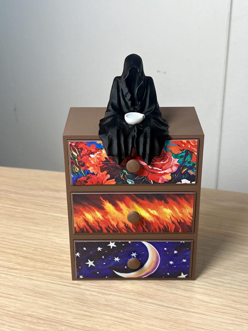 Puede incluir: Un cofre decorativo marr&oacute;n de tres cajones con frentes de cajones florales, de fuego y celestiales. Una figura negra con capucha con un cuenco blanco se sienta encima del cofre. Los cajones tienen pomos marrones.