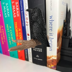 May include: A black raven bookend perched on a brown branch, holding a stack of colorful books. The bookend is next to a black castle-shaped bookend. The books have titles like "A Court of Thorns and Roses".
