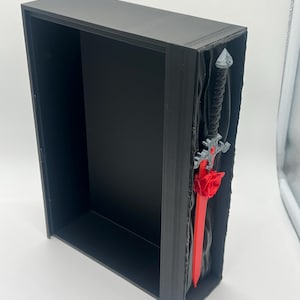 May include: A black display box with a decorative sword. The sword has a red blade and a black handle. The hilt is silver and the sword is mounted in a textured, dark gray background.