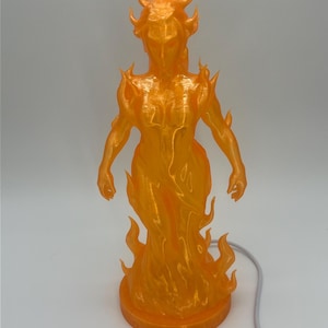 May include: An orange, flame-like sculpture of a humanoid figure. The figure has a flowing form with flames emanating from the base, head, and shoulders. The sculpture is on a round base and has a gray cord.