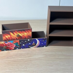 Puede incluir: Un conjunto de cajas de almacenamiento decorativas de madera y una peque&ntilde;a estanter&iacute;a. Las cajas presentan frentes de caj&oacute;n con dise&ntilde;os coloridos: un estampado floral, llamas y un cielo nocturno con una luna creciente. La estanter&iacute;a tiene tres compartimentos abiertos.