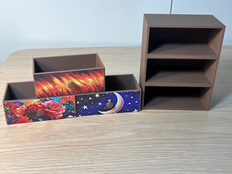 Puede incluir: Un conjunto de cajas de almacenamiento decorativas de madera y una peque&ntilde;a estanter&iacute;a. Las cajas presentan frentes de caj&oacute;n con dise&ntilde;os coloridos: un estampado floral, llamas y un cielo nocturno con una luna creciente. La estanter&iacute;a tiene tres compartimentos abiertos.