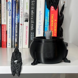 Puede incluir: Un caldero negro con tres patas, una mano gris haciendo la peineta y una peque&ntilde;a figura de una figura encapuchada. Hay libros en el fondo. Los objetos est&aacute;n sobre una estanter&iacute;a blanca.