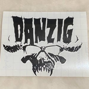 May include: Black vinyl sticker featuring the word "DANZIG" in a bold, gothic font above a stylized skull graphic. The skull has sharp teeth and a detailed design, creating a striking visual.