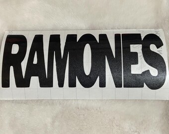 May include: Black vinyl sticker with the word "RAMONES" in a bold, sans-serif font. The letters are thick and evenly spaced, with a slight curve to the top of the letters. The sticker is rectangular and has a grid pattern on the backing.