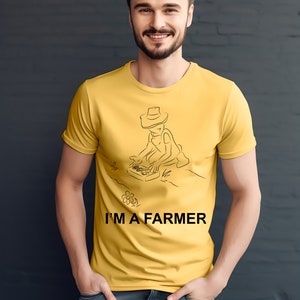 以下が含まれることがあります： マスタードイエローのTシャツ。黒い線画で農夫が描かれ、テキスト「I'M A FARMER」がプリントされています。このTシャツは、濃い髪とひげを生やし、ブルージーンズを履いた人が着用しています。