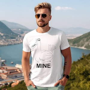 Puede incluir: Camiseta blanca con un dibujo lineal negro de un pato con gorra y un mando a distancia. La palabra "MINE" está impresa debajo del pato. La camiseta la lleva una persona.