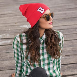 May include: A red beanie with a white "B" logo, paired with sunglasses and a green and white plaid shirt. The person is wearing dark gray jeans. The outfit is casual and stylish.