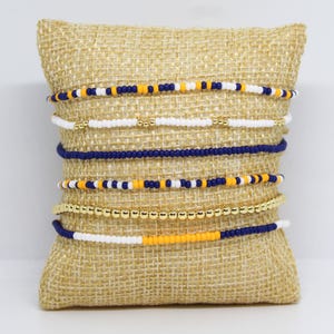 May include: A collection of beaded bracelets in various designs, displayed on a burlap pillow. The bracelets feature combinations of white, navy blue, gold, and yellow beads. The bracelets are a mix of solid colors and patterned designs.