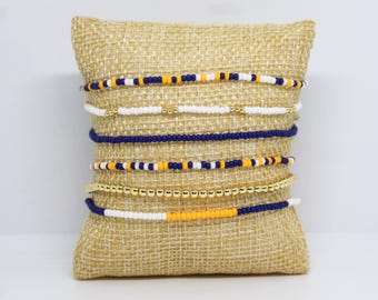 May include: A collection of beaded bracelets in various designs, displayed on a burlap pillow. The bracelets feature combinations of white, navy blue, gold, and yellow beads. The bracelets are a mix of solid colors and patterned designs.