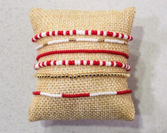 Puede incluir: Un conjunto de cinco pulseras de cuentas sobre un fondo de arpillera. Las pulseras son rojas y blancas, con algunas cuentas doradas.