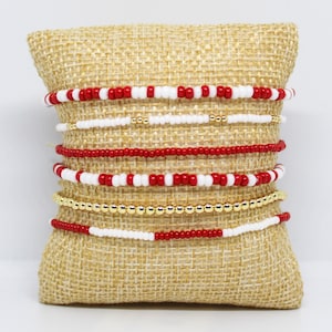 May include: A collection of beaded bracelets in red, white, and gold. The bracelets are displayed on a burlap pillow. The bracelets feature small beads in various patterns, including solid colors and alternating color combinations.