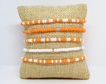 May include: A selection of beaded bracelets in orange and white are presented on a burlap cushion. The bracelets are composed of small, round beads. The bracelets feature solid orange and alternating orange and white bead patterns.