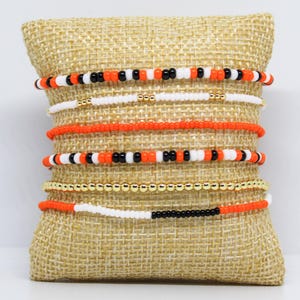 May include: A collection of beaded bracelets in various designs, displayed on a burlap pillow. The bracelets feature combinations of orange, white, black, and gold beads. Some bracelets have gold ball beads.