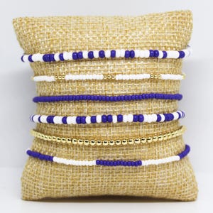May include: A collection of beaded bracelets in various designs and colors. The bracelets are displayed on a burlap pillow. The bracelets feature white, blue, and gold beads. Some bracelets have alternating colors, while others are solid.