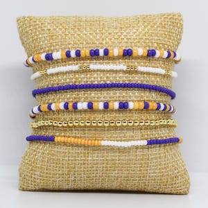 May include: A collection of beaded bracelets in various colors, including purple, gold, white, and orange. The bracelets are displayed on a burlap pillow. The beads are small and uniform in size, creating a delicate and stylish look.