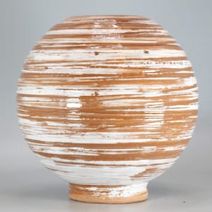 Pode incluir: Um vaso de cerâmica esférico com um padrão horizontal de riscas alternadas de esmalte branco e castanho terra. A superfície tem um aspeto texturado aplicado à mão, com uma base circular estreita e uma pequena abertura na parte superior.