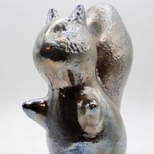 May include: A ceramic squirrel figurine with a metallic, iridescent finish. The sculpture has a textured surface, with a dark base and highlights of blue and silver. The squirrel is posed upright, with its tail curved.