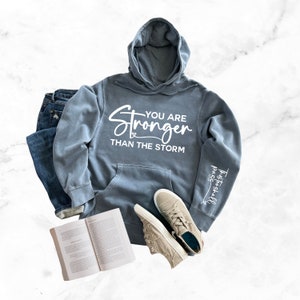 May include: A blue hooded sweatshirt with the text "You are Stronger Than The Storm" and the text "This Shall Pass" on the sleeve.