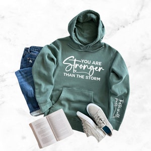 May include: A green hooded sweatshirt with the text "You are Stronger than the storm" printed on the front and the text "Trust you shall pass" printed on the sleeve.