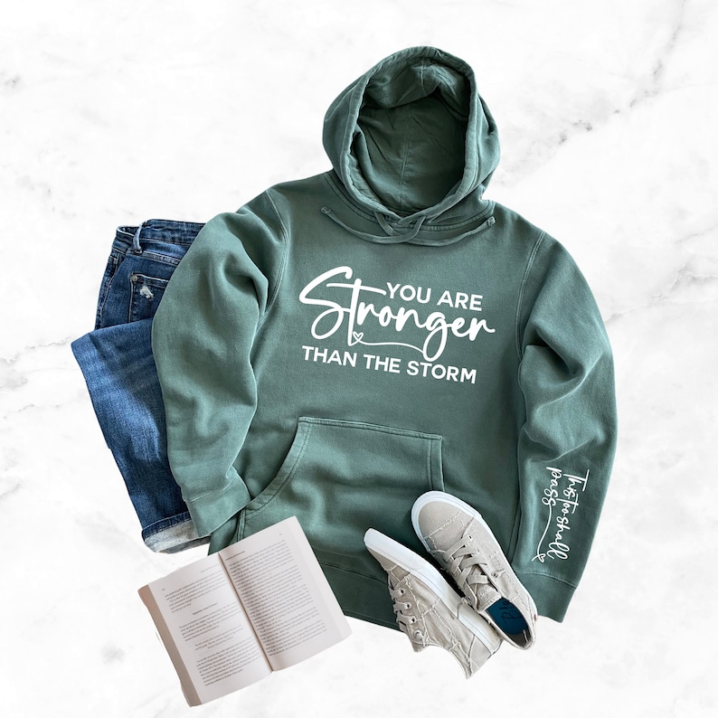 May include: A green hooded sweatshirt with the text "You are Stronger than the storm" printed on the front and the text "Trust you shall pass" printed on the sleeve.