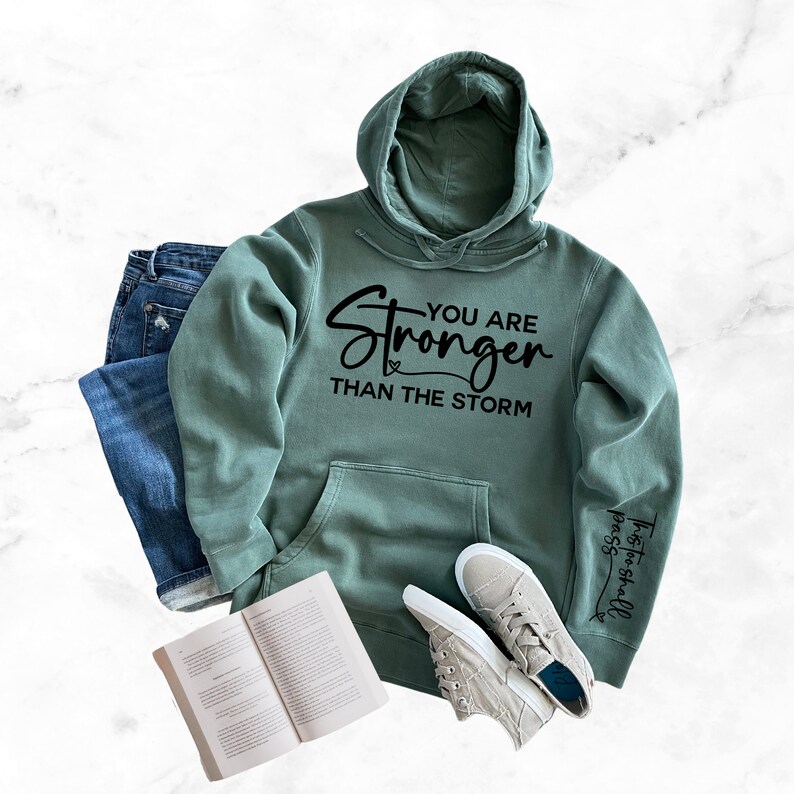 May include: A green hooded sweatshirt with the text "You are Stronger Than The Storm" and a small heart on the front. The sleeve has the text "This is the day the Lord has made, let us rejoice and be glad in it."