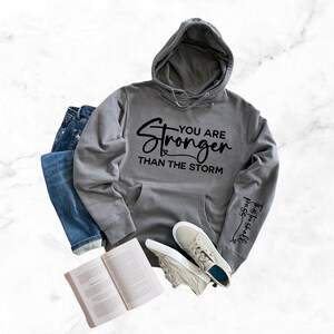 May include: Gray hooded sweatshirt with the text "You are Stronger Than The Storm" printed on the front and the text "This too shall pass" printed on the sleeve.