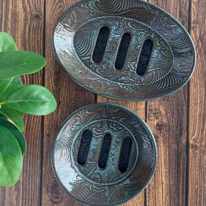 May include: Two ceramic soap dishes with a wave pattern and three drainage slots. The dishes are oval and round, with a teal and brown glaze. The soap dishes are on a wooden surface.