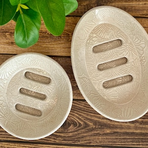 May include: Two off-white ceramic soap dishes with floral patterns. One is oval, the other is round. Each has three rectangular drainage slots. The dishes are on a wooden surface with green leaves in the background.