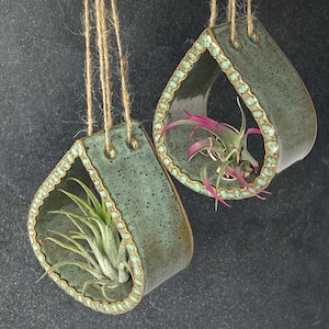 May include: Two teal green ceramic hanging planters shaped like teardrops with gold accents. Each planter has a small air plant inside.