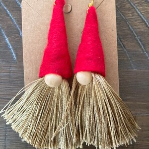 May include: A pair of red felt gnome earrings with gold tassel beards. The gnomes have wooden noses and gold earring hooks.