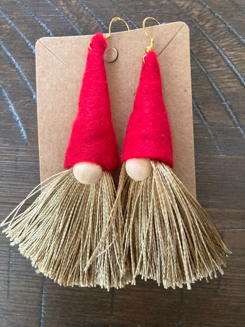 May include: A pair of red felt gnome earrings with gold tassel beards. The gnomes have wooden noses and gold earring hooks.