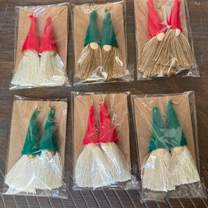 May include: Six pairs of gnome-themed tassel earrings. Each pair features a gnome with a red or green hat and a white beard. The earrings are packaged on individual cards.