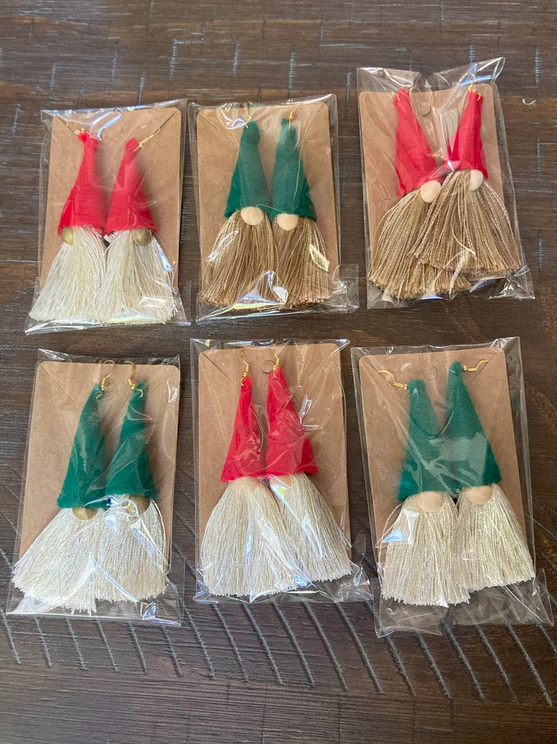 May include: Six pairs of gnome-themed tassel earrings. Each pair features a gnome with a red or green hat and a white beard. The earrings are packaged on individual cards.