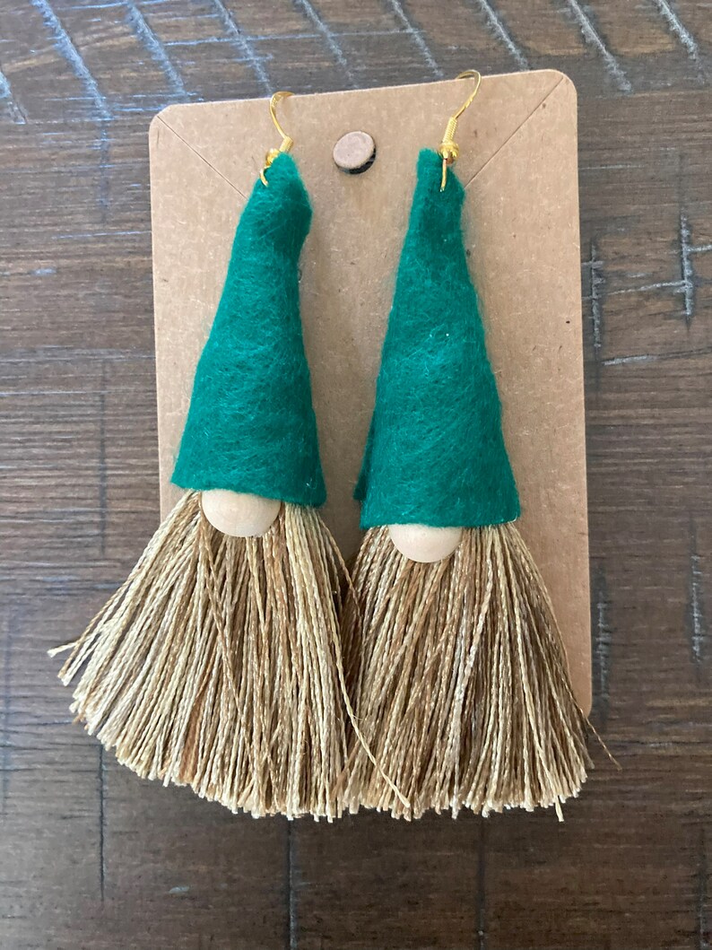 May include: A pair of green felt gnome earrings with brown yarn tassels. The earrings have wooden beads for the gnome's nose.