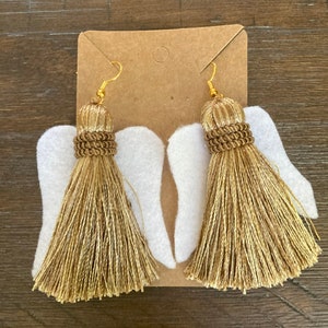 May include: A pair of gold tassel earrings with white felt angel wings. The earrings have gold hooks and are attached to the wings.
