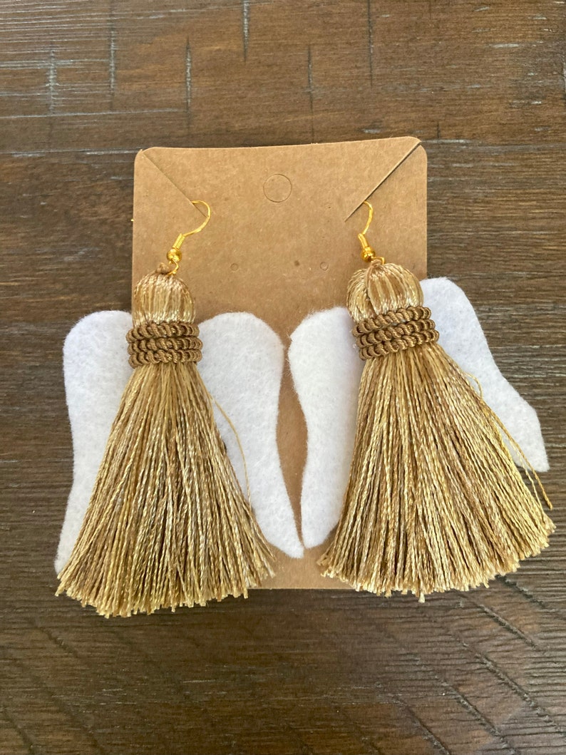 May include: A pair of gold tassel earrings with white felt angel wings. The earrings have gold hooks and are attached to the wings.